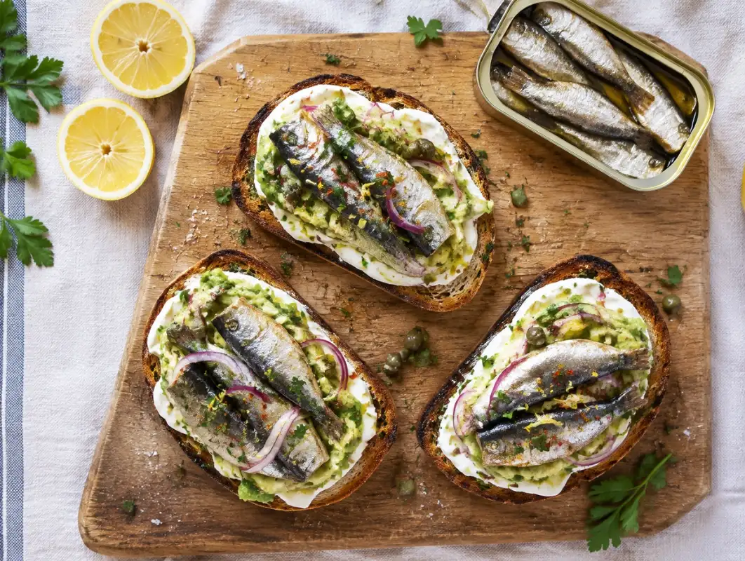 Overhead view of sardine toast on grilled sourdough with lemon zest, red onion, capers and parsley